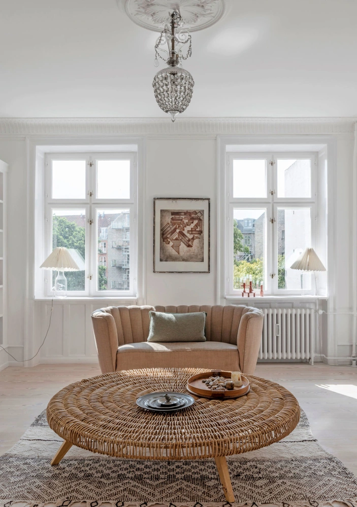Компактная софа с округлой спинкой и круглый стол из ротанга создают гармоничное оформление мягкой зоны гостиной. Рельефный рисунок на спинке софы продолжает ся в плиссерованных абажурах настольных ламп на подоконнике. | Источник: Питер Крагбалле и Кристоффер Турелл